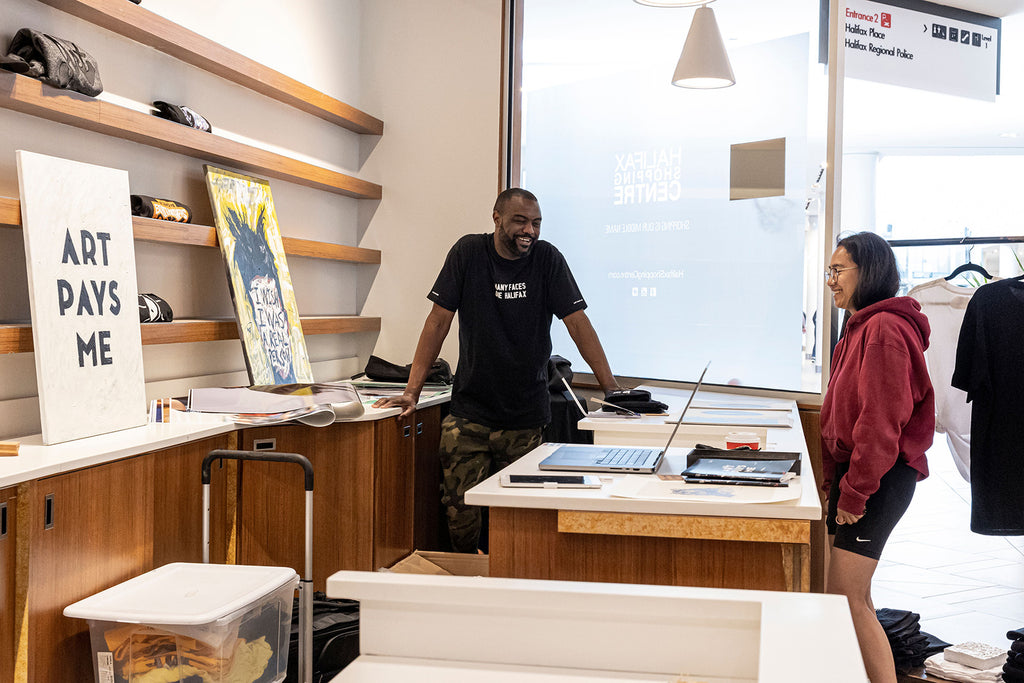 Duane talks to a customer while wearing the Many Faces One Halifax Tee at Halifax Shopping Centre during an Art Pays Me and HaliTube pop-up event. Photo by Emma Lomas.