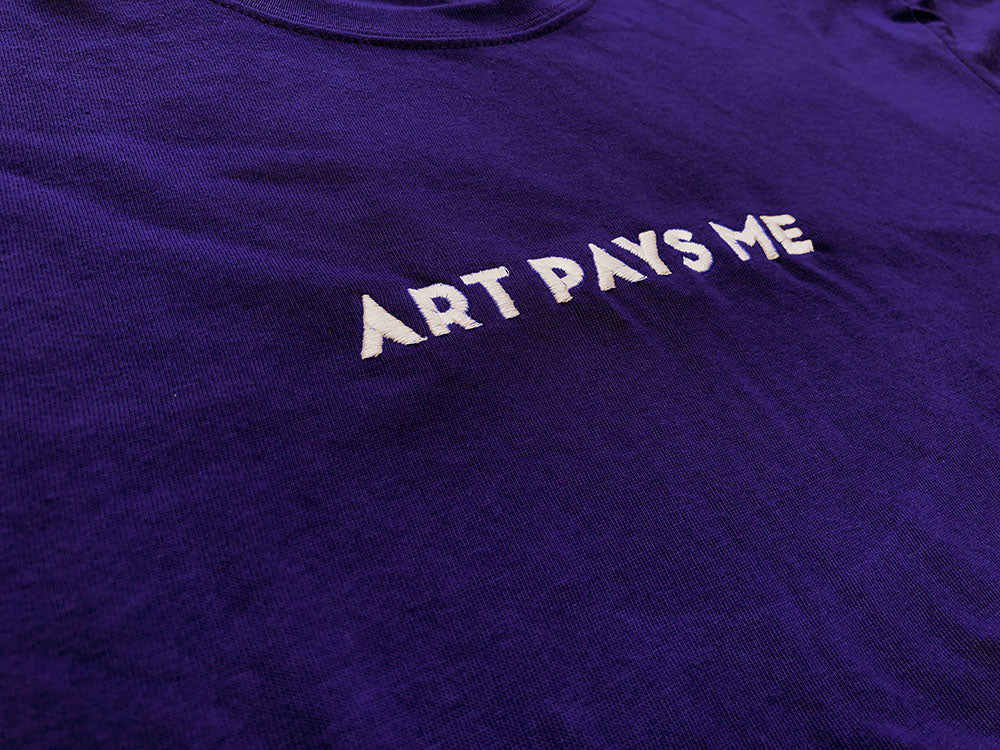 Purple 50/50 ring spun poly/cotton t-shirt with Art Pays Me embroidered on the chest in white thread.